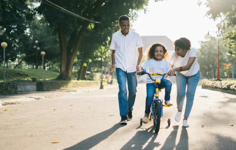 An African-American family, dad, mom, and young child, play outside on the street. The parents are helping the child learn how to ride a bike. They are all wearing white t-shirts and blue jeans