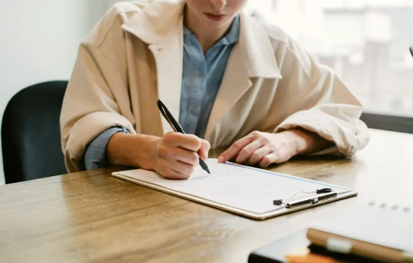 A photo of a woman filling out a form at a wooden desk. Bright light is coming through the window behind her.