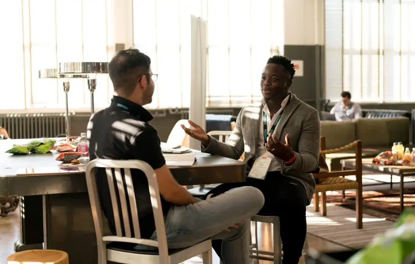 Two men are sitting at a table having a friendly conversation or interview. There are a lot of bright orange tones in the photo.