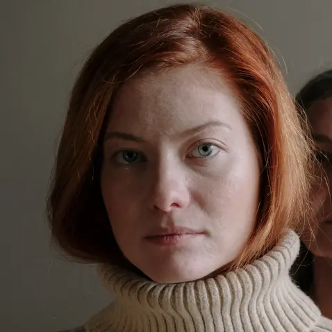 A gaunt, middle aged Caucasian woman with short red hair wearing a beige turtleneck. She looks at the camera with a grim expression.