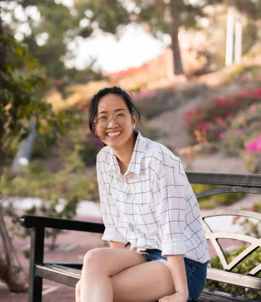 A professional picture of Shannon Lee sitting on a park bench and smiling on a sunny day. She has her hair in a pony tail and is wearing a white checkered shirt and jean shorts.